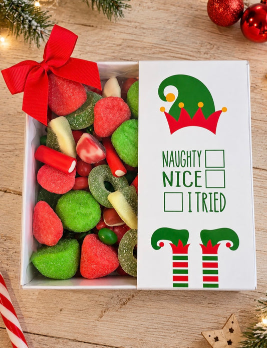 A festive Christmas gift box filled with assorted holiday candies, featuring a white card on the right with an elf hat and the text 'NAUGHTY NICE I TRIED' along with two unchecked boxes. The box is decorated with a large red bow and sits on a wooden surface with Christmas decorations.