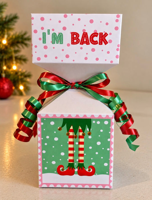 A festive Christmas gift box with an open top lid that reads 'I'm BACK' in green and red lettering. The box is decorated with pink polka dots, a green front panel featuring elf legs, and a red and green ribbon bow with curled ribbons, set against a softly blurred holiday background.