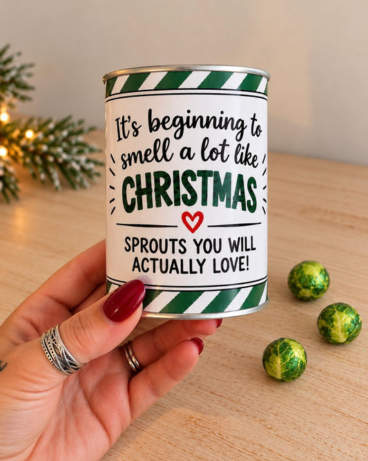 A hand holds a tin of Brussels sprouts with a humorous Christmas-themed label. The label reads 'It's beginning to smell a lot like CHRISTMAS' and 'SPROUTS YOU WILL ACTUALLY LOVE!' with green and white striped borders.